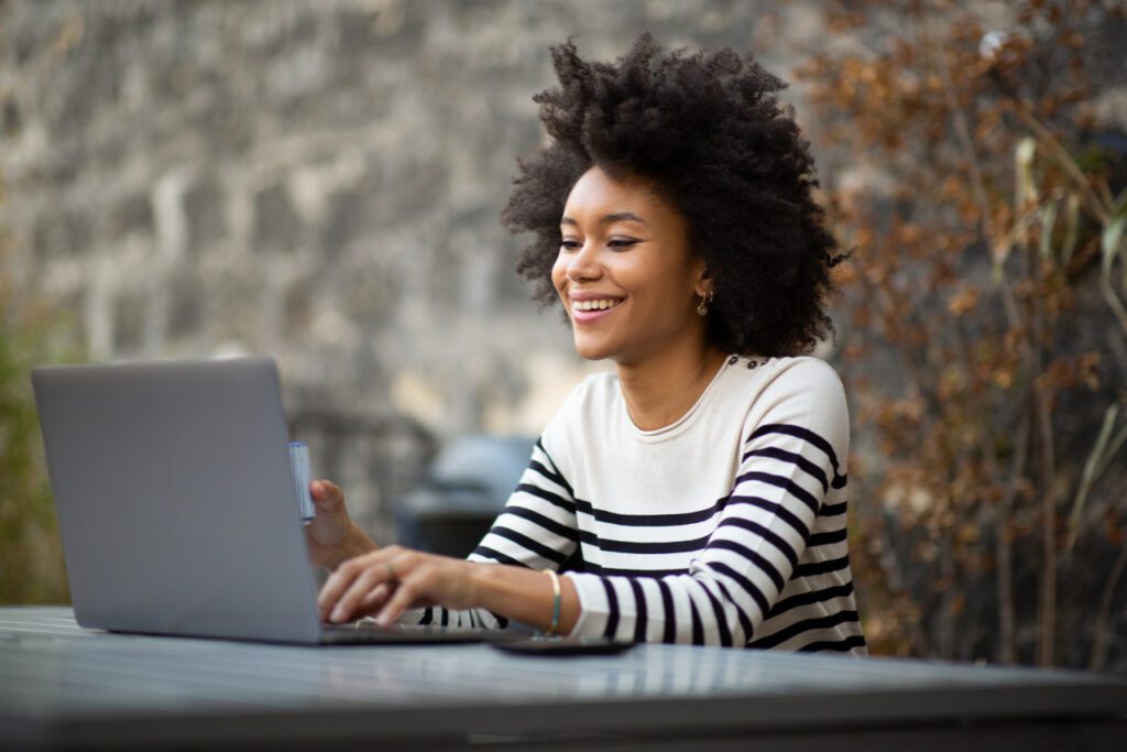 Image of a woman sitting outside and smiling while working on her laptop.