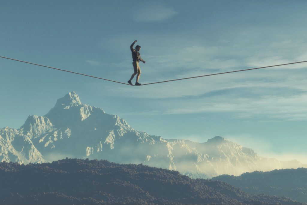 Image of a man balancing on a tight rope overlooking a vast mountain. 