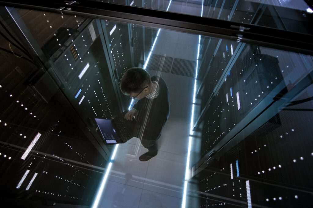 An image of a man standing in a server room using his laptop to upload all of his commission data. 