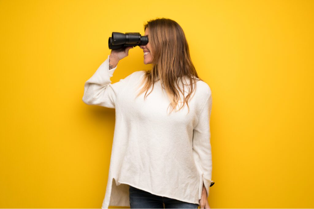 An image of a woman standing in front of a yellow screen looking to her right through binoculars. 